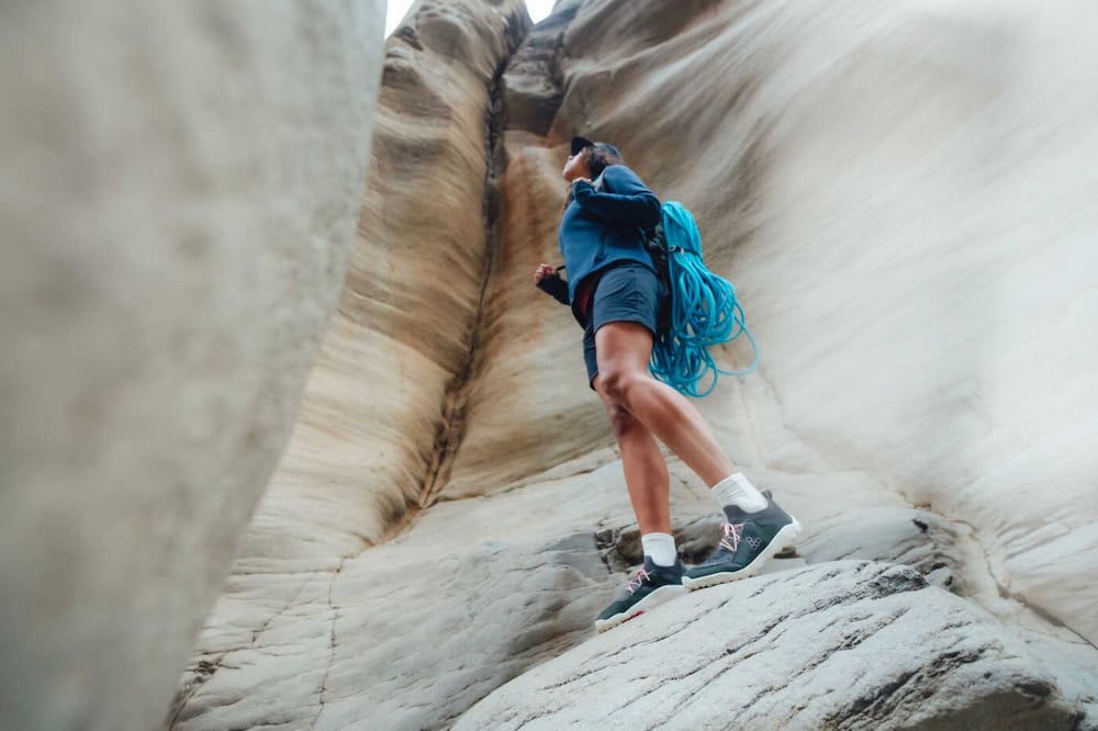 A climber standing on a mountain wearing shorts and thin soled blue shoes, carrying a hank of rope over her back and looking upwards