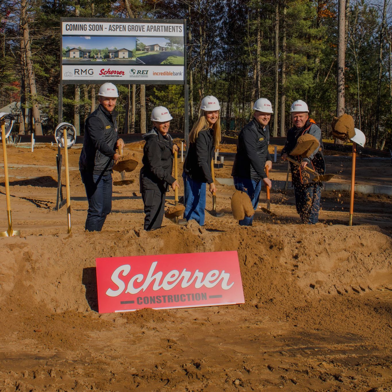 Five people in hard hats turning soil at a construction groundbreaking site in Oneida County, Wisconsin, with trees and a project sign in the background.