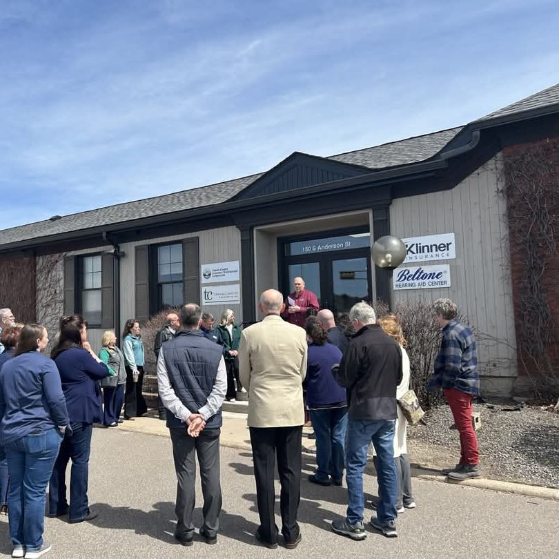 A group of people is gathered in front of a building with blue skies above, attending a ribbon cutting ceremony. The building has signage and a person is speaking on the front steps.