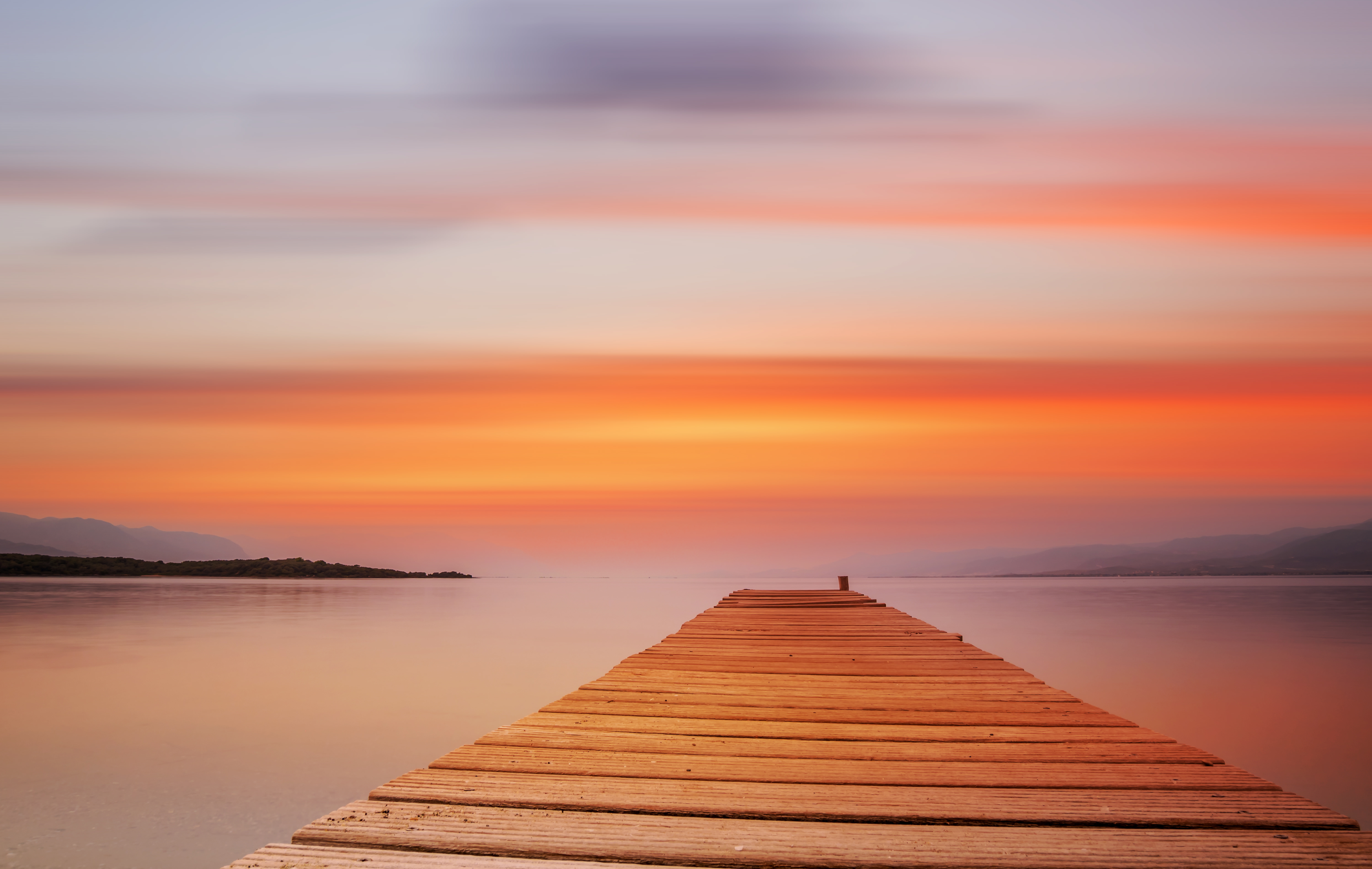 A serene view of a wooden pontoon extending into calm waters against a vibrant sunset sky on Evia Island, Greece