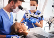 Zahnbehandlung: Ein Zahnarzt schaut durch ein Mikroskop und behandelt eine Patientin. Daneben sitzt eine Zahnarzthelferin.