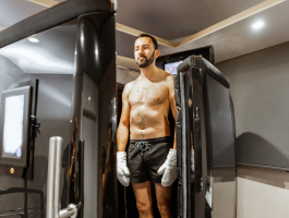Ein Mann in Shorts und Handschuhen steht in einer Kryokammer in einem modernen Wellness-Zentrum. Er bereitet sich auf eine Kältetherapie vor, umgeben von futuristischer Ausrüstung und gedämpfter Beleuchtung.