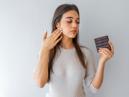 Eine Frau, die in der einen Hand eine Tafel Schokolade hält und mit der anderen auf das Rosacea auf ihrer Wange zeigt.