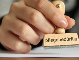 Nahaufnahme einer Hand mit Holzstempel „pflegebedürftig“, der auf ein Blatt Papier gedrückt wird – Symbol für amtliche Anerkennung von Pflegebedürftigkeit. ChatGPT fragen