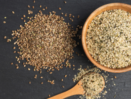 Ungeschälte Hanfsamen und geschälte Hanfkerne in einer Holzschale sowie auf einem Löffel, arrangiert auf dunkler Steinoberfläche – Darstellung verschiedener Formen von Hanfsamen für Ernährung und Lebensmittelverarbeitung.