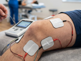 Patient liegt in einer Physiotherapiepraxis auf einer Behandlungsliege, während Elektroden zur Iontophorese am Knie angebracht sind. Die Stromtherapie dient der gezielten Behandlung von Schmerzen, Entzündungen oder Gelenkbeschwerden.