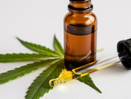Braune CBD-Ölflasche mit Pipette und goldfarbenem Öl auf grünen Hanfblättern vor hellem Hintergrund.