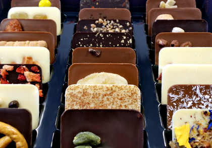 Eine Schachtel mit verschiedenen rechteckigen Schokoladenstücken, die jeweils mit unterschiedlichen Toppings wie Nüssen, getrockneten Früchten, Streuseln, Keksstücken und Blüten verziert sind.