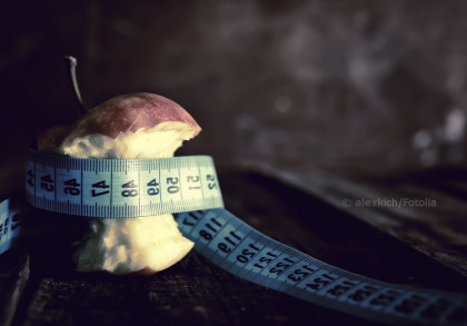 Ein angebissener Apfel, der von einem blauen Maßband umwickelt ist, liegt auf einer dunklen Oberfläche vor einem unscharfen Hintergrund.