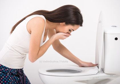 Eine Frau mit langen braunen Haaren und einem weißen Tanktop beugt sich über eine Toilette und hält sich den Mund, als ob sie sich übergeben müsste.