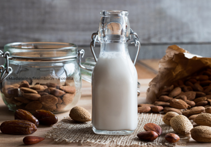 Ein Glasflasche mit Mandelmilch steht auf einem Holztisch. Im Hintergrund befindet sich ein Glasgefäß mit Mandeln sowie ein geöffneter Papiersack mit weiteren Mandeln. Um die Flasche herum liegen einige Datteln und rohe Mandeln auf dem Tisch verstreut.