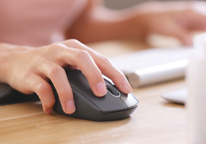 Eine Hand bewegt eine Computermaus auf einem Holztisch. Im Hintergrund sind unscharf eine Tastatur und andere Objekte zu sehen.