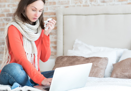 Eine Frau sitzt auf einem Bett mit einem Laptop auf ihrem Schoß. Sie trägt einen weißen Schal und einen orangefarbenen Pullover. In der rechten Hand hält sie ein Taschentuch, als ob sie sich erkältet hätte.