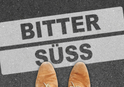 Die Bildbeschreibung zeigt zwei Straßenschilder auf einem Asphaltuntergrund. Das obere Schild weist nach links und trägt die Aufschrift "BITTER", das untere Schild weist nach rechts und trägt die Aufschrift "SÜSS".