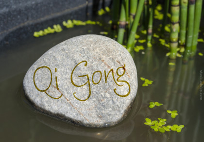 Ein großer, glatter Stein liegt im Wasser, auf dessen Oberfläche das Wort "Qi Gong" geschrieben ist. Im Hintergrund sind Bambusrohre und schwimmende Pflanzenblätter zu sehen. Die Umgebung vermittelt eine ruhige und friedliche Atmosphäre.