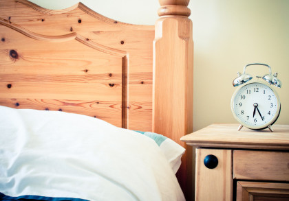 Holzbett mit Kopfteil und weißer Bettwäsche daneben ein hölzerner Nachttisch mit einem silbernen Wecker.