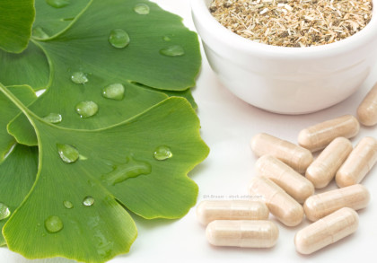 Nahaufnahme von grünen Ginkgo-Blättern mit Wassertropfen, einer weißen Schüssel mit getrockneten Kräutern und mehreren Kapseln mit Kräuterinhalt auf einer weißen Oberfläche.