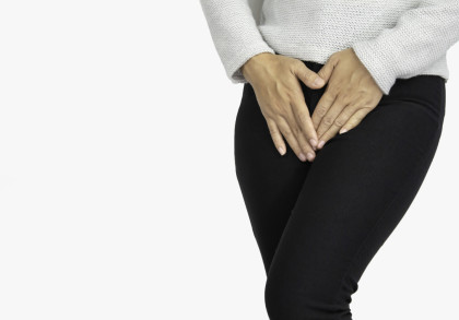 Eine Person in einem weißen Pullover und schwarzen Hosen hält beide Hände in der Nähe des Unterbauchs.