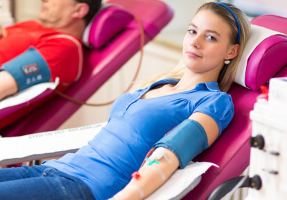 Eine junge Frau sitzt auf einem medizinischen Liegestuhl und spendet Blut. Im Hintergrund ein weiterer Blutspender. Beide haben Blutdruckmanschetten am Arm und sind mit medizinischen Geräten verbunden.