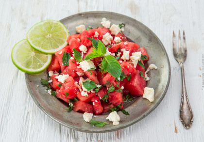 Ein Teller mit Wassermelonen-Feta-Salat, garniert mit frischer Minze und zwei Limettenscheiben auf der Seite. Rechts vom Teller liegt eine antike Gabel auf einem weißen Holztisch.