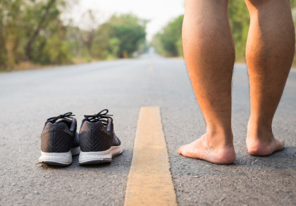 Ein Mann steht barfuß auf der Straße neben einem Paar schwarzer Sportschuhe. Die Straße führt in die Ferne und ist von Bäumen gesäumt.
