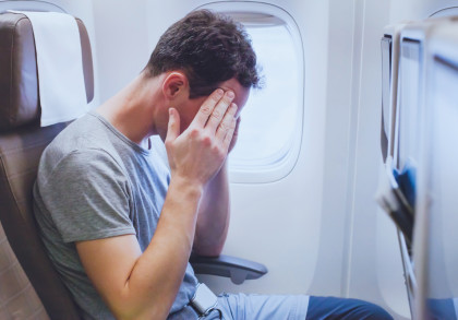 Ein Mann sitzt in einem Flugzeugsitz und hat sein Gesicht in seine Hände gelegt. Er trägt ein graues T-Shirt und wirkt besorgt oder gestresst. Im Hintergrund sieht man ein Flugzeugfenster.