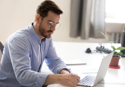 Ein Mann mit Brille und hellblauem Hemd sitzt an einem Schreibtisch und arbeitet konzentriert an einem Laptop. Es gibt einige Dokumente auf dem Tisch und eine Pflanze im Hintergrund.