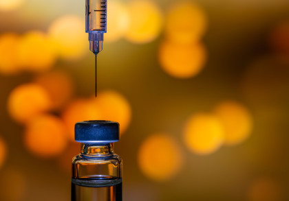 Nahaufnahme einer Spritze über einem Fläschchen mit orangefarbenen Bokeh-Lichtern im Hintergrund. Die Nadel der Spritze ist nur wenige Millimeter über einer Gummikappe des Fläschchens positioniert.