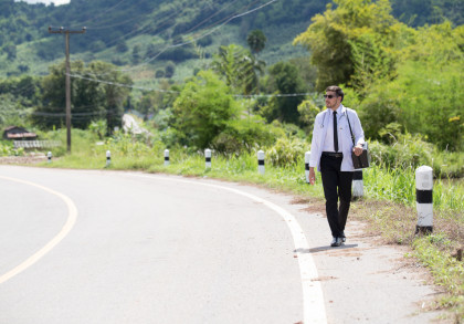 Ein Mann im Arztkittel mit einer Tasche und einer Sonnenbrille geht am Straßenrand einer ländlichen Straße entlang, umgeben von grüner Landschaft. Im Hintergrund sind Hügel und Bäume zu sehen. Es ist ein sonniger Tag mit klarem Himmel.