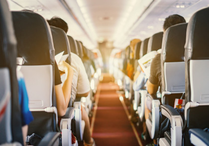 Innenansicht eines Flugzeugs mit Reihen von Passagieren, die nach vorne blicken. Einige Passagiere lesen oder halten Gegenstände in der Hand. Die Gänge zwischen den Sitzen sind deutlich sichtbar.