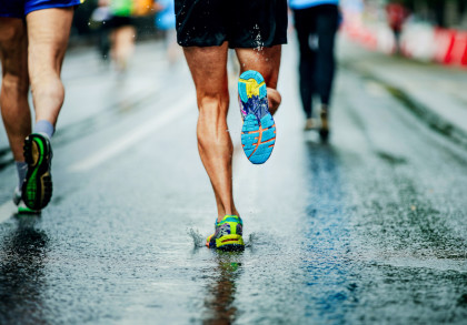 Nahaufnahme von Läufern in bunten Laufschuhen, die bei nassem Wetter auf einer Straße rennen. Wassertropfen spritzen von den Füßen auf den nassen Boden.