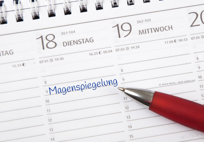 Ein Kalender zeigt den Dienstag, den 18., und Mittwoch, den 19., wobei am 18. eine Notiz „Magenspiegelung“ mit blauer Tinte eingetragen ist. Ein roter Kugelschreiber liegt auf dem Kalender.