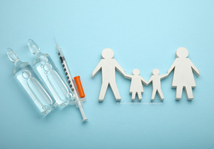 Zwei Ampullen mit Flüssigkeit und eine Spritze liegen auf blauem Hintergrund. Neben den Medikamenten sind Holzfiguren, die eine Familie darstellen, abgebildet. Die Familie besteht aus zwei Erwachsenen und zwei Kindern, die sich an den Händen halten.