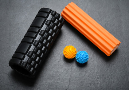 Bildbeschreibung in Deutsch: Zwei Massagerollen, eine schwarze und eine orangefarbene, liegen auf einem dunklen Hintergrund. Neben ihnen liegen zwei stachelige Massagebälle, einer gelb und einer blau.