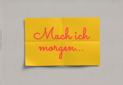 Das Bild zeigt ein gelbes Blatt Papier mit dem handgeschriebenen Text "Mach ich morgen..." in pinker Farbe. Das Blatt ist leicht gefaltet und vor einem grauen Hintergrund platziert.