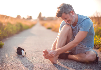 Ein Mann sitzt auf einem ländlichen Weg und hält seinen Fuß mit einem schmerzverzerrten Gesichtsausdruck. Sein linker Schuh liegt neben ihm auf dem Boden. Es ist ein sonniger Tag, und der Weg ist von Gras und Pflanzen gesäumt.