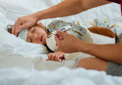 Eine Hand hält ein Thermometer, während die andere Hand die Stirn eines schlafenden Kindes fühlt, das im Bett liegt und ein Kühlkissen auf der Stirn hat. Das Kind trägt einen bunten Schlafanzug mit Tiermotiven.