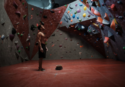 Ein muskulöser Mann steht in einer Kletterhalle mit rot-grau gestreiften Kletterwänden und farbigen Griffen. Er trägt schwarze Shorts und steht auf einem roten Bodenbelag, während ein Kreidebeutel zwischen seinen Füßen liegt.