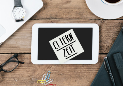 Auf einem Holztisch liegen ein Laptop, eine Armbanduhr, eine Tasse Kaffee, ein Taschenrechner, eine Brille, bunte Büroklammern, ein Notizbuch mit Stift und ein Tablet mit einem Klebezettel darauf. Auf dem Zettel steht in Großbuchstaben "ELTERNZEIT".