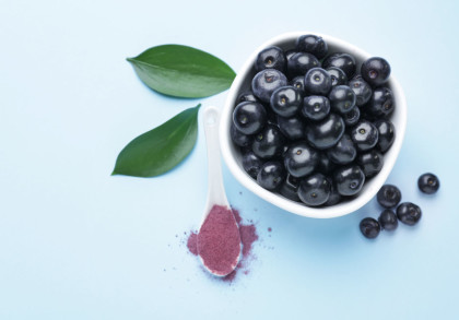 Eine weiße Schüssel gefüllt mit Acai-Beeren, daneben ein weißer Löffel mit Acai-Pulver auf einem hellblauen Hintergrund. Zwei grüne Blätter liegen neben der Schüssel.