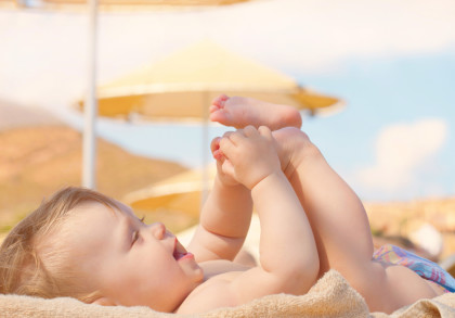 Ein lächelndes Baby, das auf einem Handtuch liegt und mit seinen Füßen spielt. Im Hintergrund sind Sonnenschirme und ein heller, wolkenloser Himmel zu sehen.