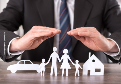 Ein Mann in Anzug und Krawatte hält seine Hände schützend über Papiersilhouetten einer Familie mit zwei Kindern, einem Haus und einem Auto auf einem Tisch.
