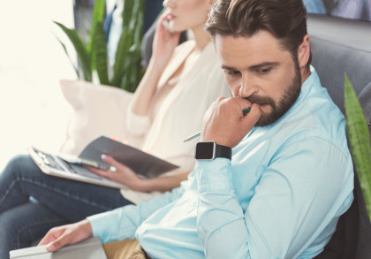 Ein nachdenklich aussehender Mann sitzt auf einem Sofa mit einer Smartwatch am Handgelenk und einem Stift in der Hand. Neben ihm sitzt eine Frau mit einem Laptop und einem Notizbuch, sie telefoniert. Im Hintergrund sind Pflanzen sichtbar.