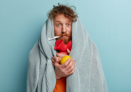Ein Mann mit zerzaustem Haar und rotem Bart ist in eine graue Decke eingehüllt. Er hat ein Thermometer im Mund und hält ein rotes Stethoskop sowie eine Zitrone in seinen Händen. Der Hintergrund ist hellblau.