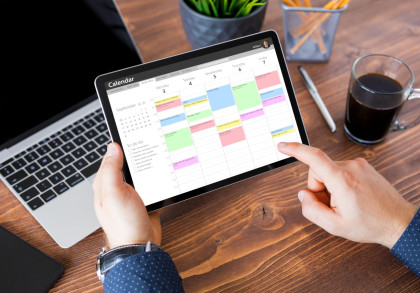 Eine Person benutzt ein Tablet, das einen digitalen Kalender mit farblich markierten Terminen für die Woche anzeigt. Auf dem Holztisch befinden sich ein Laptop, ein Smartphone, eine Tasse Kaffee, ein Stifthalter mit Stiften und ein Kugelschreiber.