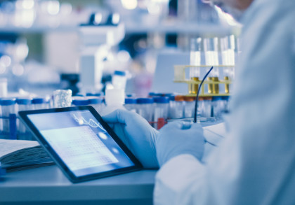 Ein Wissenschaftler in einer Laborumgebung untersucht Daten auf einem Tablet. Im Hintergrund sind mehrere Reagenzgläser, Laborgeräte und Probenbehälter zu sehen. Der Wissenschaftler trägt einen weißen Laborkittel und blaue Handschuhe.