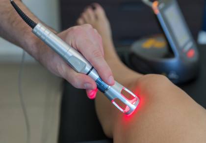 Nahaufnahme eines Physiotherapeuten, der eine Lasertherapie-Einheit auf ein Knie anwendet. Der Laser leuchtet rot und wird auf die Haut gerichtet.
