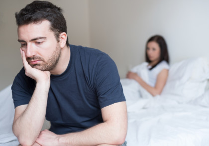 Ein Mann mit dunklem Haar und Bart sitzt nachdenklich auf einem Bett, stützt seinen Kopf auf seine Hand und schaut nach unten. Im Hintergrund sitzt eine Frau mit dunklem Haar in einem weißen Oberteil auf dem Bett, sie wirkt ebenfalls nachdenklich.