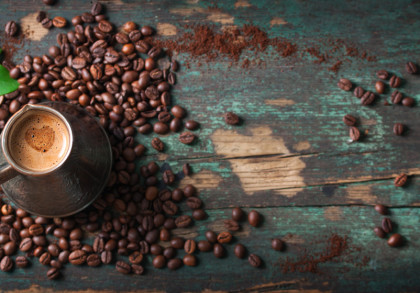 Ein traditionelles Kaffeekännchen, gefüllt mit heißem Kaffee, steht auf einem Tisch, der mit verstreuten Kaffeebohnen und gemahlenem Kaffee bedeckt ist. Daneben sind grüne Blätter zu sehen.