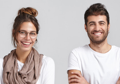 Ein Mann und eine Frau stehen vor einem grauen Hintergrund und lächeln in die Kamera. Die Frau trägt eine Brille und einen beigen Schal. Der Mann hat dunkles Haar und trägt ein weißes T-Shirt.